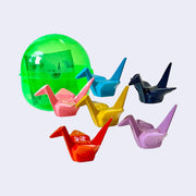 Bright green plastic capsule, positioned behind 6 different solid colored figures modeled to look like origami cranes. Colors are: pink, blue, navy, yellow, lilac, and red.