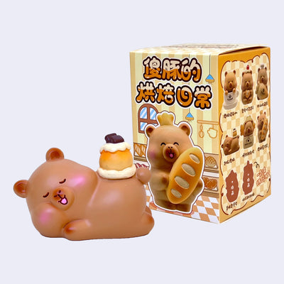 Cute capybara figurine, resting happily on the ground with a cake on its back. It is in front of its blind box packaging.