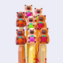 Several bright colored pens - mostly yellow, orange and green, with rubber toppers of cartoon style capybaras, sitting and holding a fruit to their stomach.