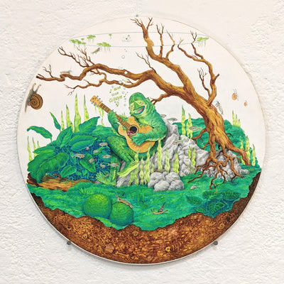 Drawing on circular panel of a Loch Ness monster creature, strumming a guitar and singing happily. It sits on a rock formation in a terrarium setting with lots of greenery and a tree branch behind it. A cross section of the underground is visible, with some fish bones visible. 