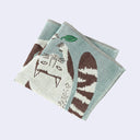 Teal blue square handkerchief towel with an illustration of a white cat with brown stripes and spots, playing with a floating green leaf. It is next to a stylized tree, the same size as itself.
