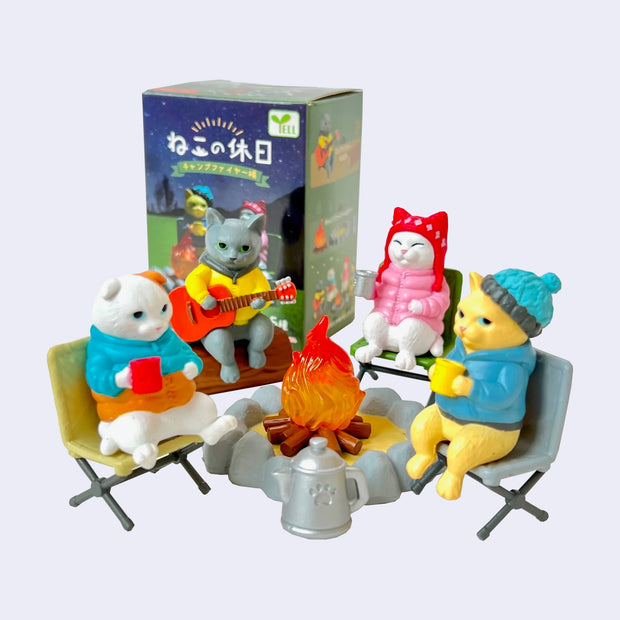 Group of small plastic cats dressed in warm headwear and jackets, gathered around a small campfire either drinking from mugs or playing the guitar.