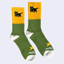 Pair of socks - olive green, yellow and white with small graphics of a black cat carry its small kitten in its mouth.
