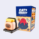 Small plastic Siamese cat figurine, with a boxy body and positioned like rice for tamago sushi. It's wrapped with a black seaweed strip and sits on a black plate. It stands in front of its product packaging.