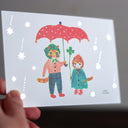 Hand holding a postcard with an illustration of 2 cats standing under an umbrella.