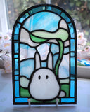 Stained glass small flat arch shaped piece, with blue glass around the border and a cute, small white bunny like character in the middle. It holds a leaf umbrella over its head.
