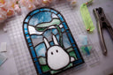 Stained glass small flat arch shaped piece, with blue glass around the border and a cute, small white bunny like character in the middle. It holds a leaf umbrella over its head.