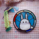 Circular stained glass hanging with a rainbow colored exterior circle, and interior blue background and a mostly white Chibi Totoro character.