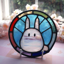 Circular stained glass hanging with a rainbow colored exterior circle, and interior blue background and a mostly white Chibi Totoro character.