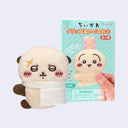 Plush toy resembling an otter with a hand holding it, next to its packaging on a light blue background.