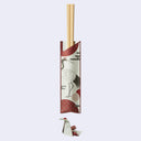 A pair of light wood chopsticks resting in a decorative paper chopstick sleeve featuring a crane illustration and red geometric patterns, shown on a plain light gray background with a small folded paper crane below.