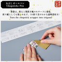 A close-up of hands folding a small origami paper airplane on a gray surface, with a printed folding instruction sheet and Japanese text above. The product appears to be a chopstick pen and origami paper set, and the image is instructional.