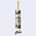 A pair of light wood chopsticks resting in a decorative paper sleeve with a Japanese-style samurai helmet and red bow illustration, shown on a pale gray background with a small matching paper accessory below.