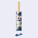 A pair of light wood chopsticks resting in a decorative blue and white chopstick sleeve with a Japanese mountain and sun motif, shown on a plain light gray background with a small separate folded paper piece below.