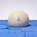 Carved wooden sculpture of a wide cream colored moon, with a flat bottom. It has several small craters and a closed eye, calm facial expression. 