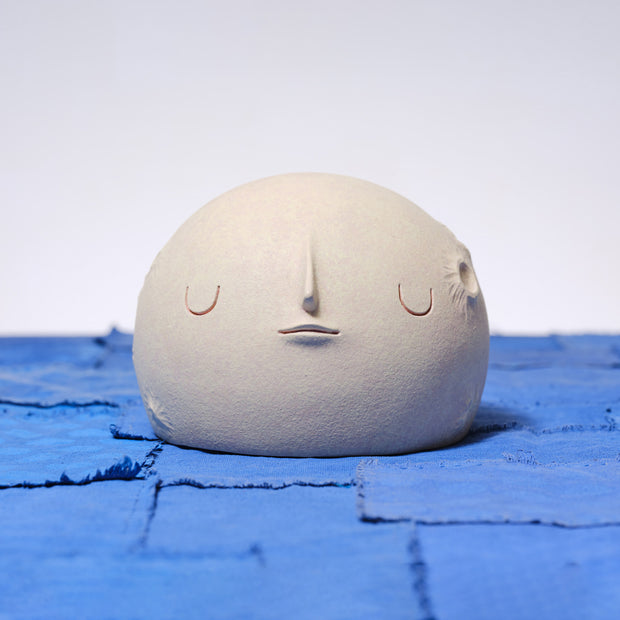 Carved wooden sculpture of a wide cream colored moon, with a flat bottom. It has several small craters and a closed eye, calm facial expression. 
