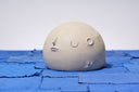 Carved wooden sculpture of a wide cream colored moon, with a flat bottom. It has several small craters and a closed eye, calm facial expression. 