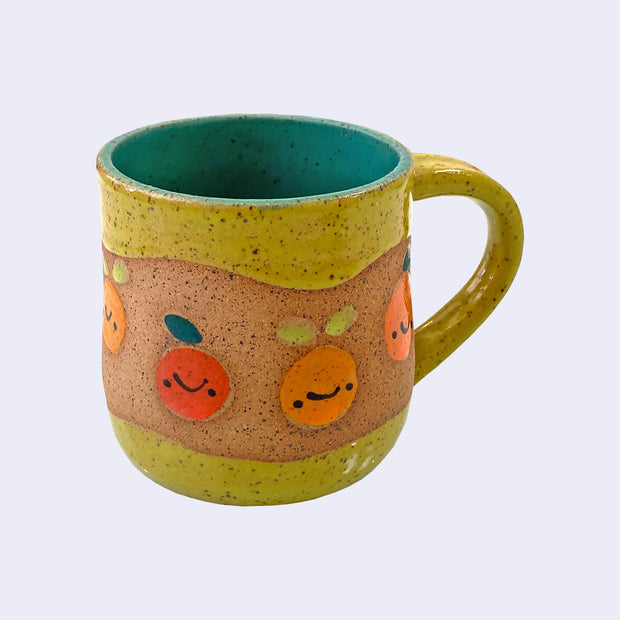 Ceramic mug with spotted finishing and an earthy brown exterior and teal blue interior. On the outside are painted on cartoon style citrus fruits, with simple expressions.