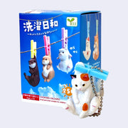 Cat figurine with a blue clothesline pin on the scruff of its back and packaging featuring animal figures on a clothesline.