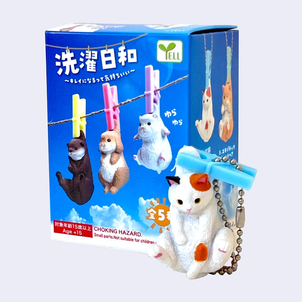 Cat figurine with a blue clothesline pin on the scruff of its back and packaging featuring animal figures on a clothesline.