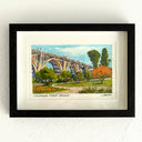 A framed artwork depicting the Colorado Street Bridge in Pasadena, featuring a colorful landscape painting with green trees, orange foliage, and a blue sky, set in a black frame with a white mat on a textured white wall.