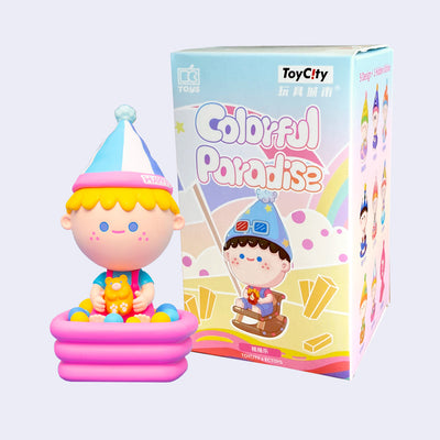 Colorful vinyl figure of a small blonde cartoon style boy sitting in an inflatable ball pit with a teddy bear. He wears a pointed cap and sits in front of its product packaging.