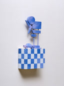 Small rectangular wood panel painted in a white and night sky blue checkered pattern. Atop of it are carved wooden sculptures: a bluish purple retro style car and several large sunset colored Americana style signs, attached to 2 tall poles.