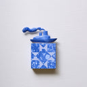 Small square wood panel painted in a blue splatter pattern with light blue patterning overlayed on top, akin to a quilt tile design. Atop of it is a carved wooden sculpture of a boat with a cloud of smoke coming out the top.