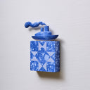 Small square wood panel painted in a blue splatter pattern with light blue patterning overlayed on top, akin to a quilt tile design. Atop of it is a carved wooden sculpture of a boat with a cloud of smoke coming out the top.