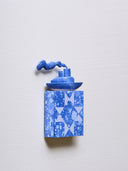 Small square wood panel painted in a blue splatter pattern with light blue patterning overlayed on top, akin to a quilt tile design. Atop of it is a carved wooden sculpture of a boat with a cloud of smoke coming out the top.