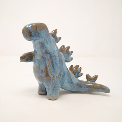 Ceramic sculpture of a Godzilla monster, with an overall smooth body shape and no distinct facial features. It has spikes running down its back and has one hand extended out slightly. Colors are blue with brown drips.