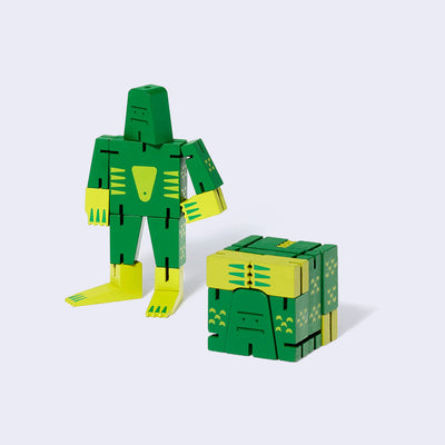 Wooden swamp monster character made of green and yellow square-like shapes, standing with one arm on its hip and the other at its side. In front is the same doll, folded into the shape of a perfect cube. 