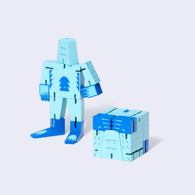 Wooden yeti character made of light and dark blue square-like shapes, standing with one arm on its hip and the other at its side. In front is the same doll, folded into the shape of a perfect cube. 