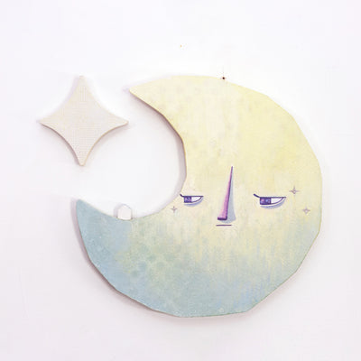 Die cut canvas sculpture of a crescent moon, with an apathetic expression. It looks off to the side at a tiny house that sits on the moon. A star hangs in the upper left corner.