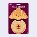 Set of 2 sponges on a backing card that reads "Daily Bread". One sponge is shaped like a croissant and the other is shaped like an everything bagel with a bite taken out of it.