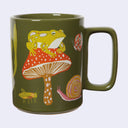 Olive green mug with an illustration of a frog sitting atop a mushroom on it, with other bugs around them both.