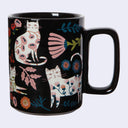 A glossy black ceramic mug decorated with whimsical white cats and colorful floral motifs, shown against a plain light gray background.