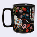 A glossy black ceramic mug decorated with whimsical white cats and colorful wildflowers, shown against a light gray background.