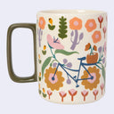 A ceramic mug with a cream-colored base decorated with a whimsical pattern of flowers, cacti, and a blue bicycle. The mug features a solid dark olive green handle and is set against a plain light gray background.