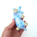 A hand holding a small blue and white spotted ceramic figurine of a dog lying on its back, with gold-painted paw pads and a white peace sign on its belly, against a plain white background.