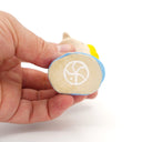 A hand holding a small ceramic figurine, showing the bottom side of the figure which features a white printed symbol on a beige base with blue and yellow accents.