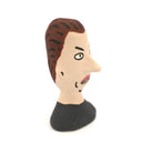 A hand-painted clay figurine of a man's head with brown hair, a beige face, black outlines, and a black shirt, shown against a plain white background.