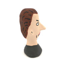 A hand-painted clay figurine of a man's head with brown hair, a beige face, black outlines, and a black shirt, shown against a plain white background.