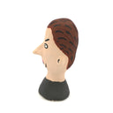A hand-painted clay figurine of a man's head with brown hair, a beige face, black outlines, and a black shirt, shown against a plain white background.