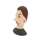 A hand-painted clay figurine of a man's head with brown hair, a beige face, black outlines, and a black shirt, shown against a plain white background.