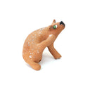 A small sculpted dog figurine in a warm tan-brown glaze with white speckles, a green eye, and a black nose, scratching behind its ear, photographed on a plain white background.