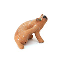 A small sculpted dog figurine in a warm tan-brown glaze with white speckles, a green eye, and a black nose, scratching behind its ear, photographed on a plain white background.