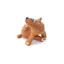 A small sculpted dog figurine in a warm tan-brown glaze with white speckles, a green eye, and a black nose, scratching behind its ear, photographed on a plain white background.