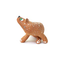 A small sculpted dog figurine in a warm tan-brown glaze with white speckles, a green eye, and a black nose, scratching behind its ear, photographed on a plain white background.