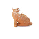 A small sculpted dog figurine in a warm tan-brown glaze with white speckles, a green eye, and a black nose, scratching behind its ear, photographed on a plain white background.